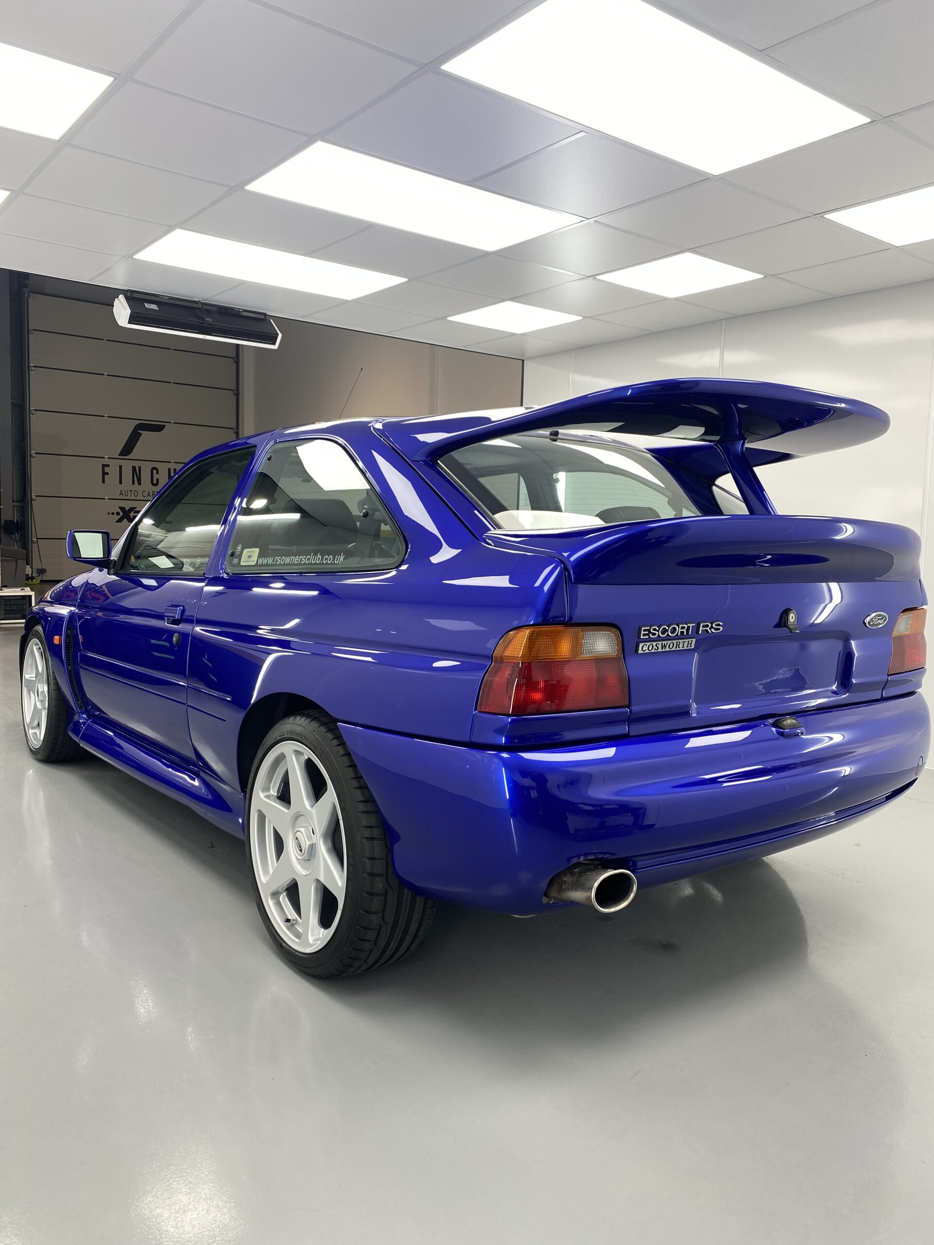 A blue ford escort rs cosworth with a large rear wing parked indoors.