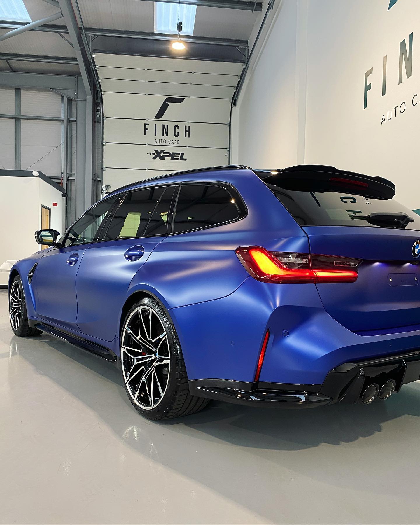 A blue bmw m3 estate parked in a showroom with focus on its rear and side profile.