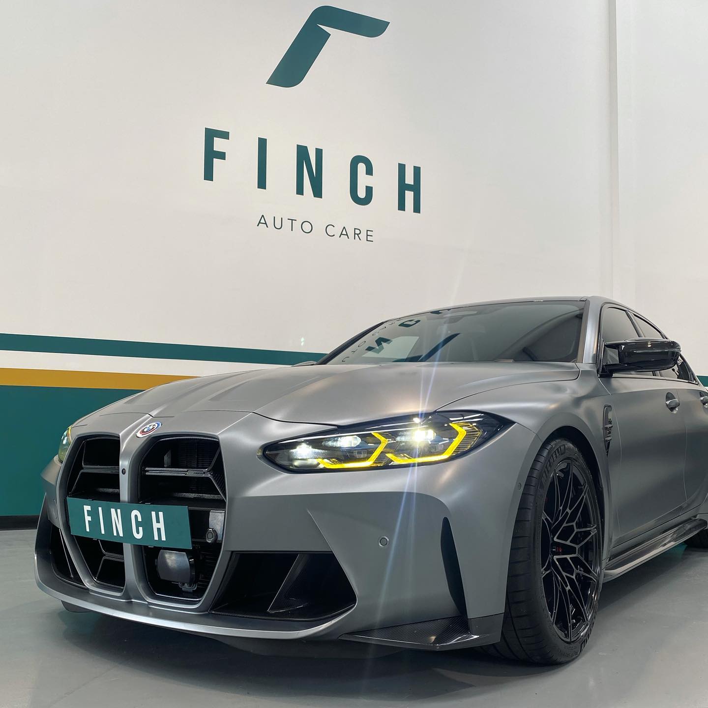 A matte grey sports car displayed in a showroom with the "finch auto care" logo on the wall behind it.